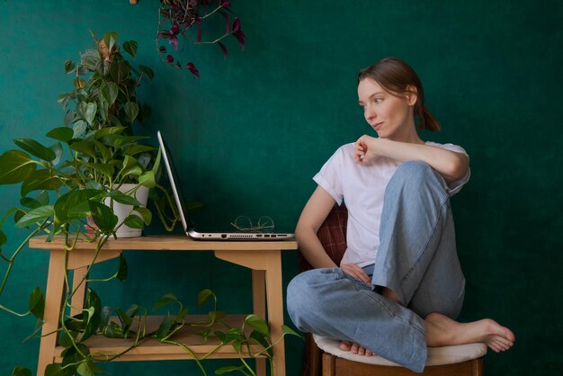 Vrolijke jonge vrouw in spijkerbroek en wit t-shirt thuis aan tafel op een zonnige dag die online of op afstand leert