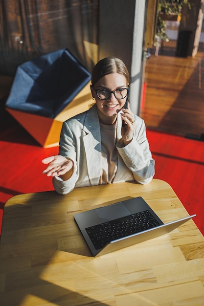 Vrolijke jonge vrouw in vrijetijdskleding met telefoongesprek zittend op de werkplek in de buurt van laptop op kantoor glimlachende vrouwelijke freelancer die naar collega belt om goed nieuws over zaken te delen