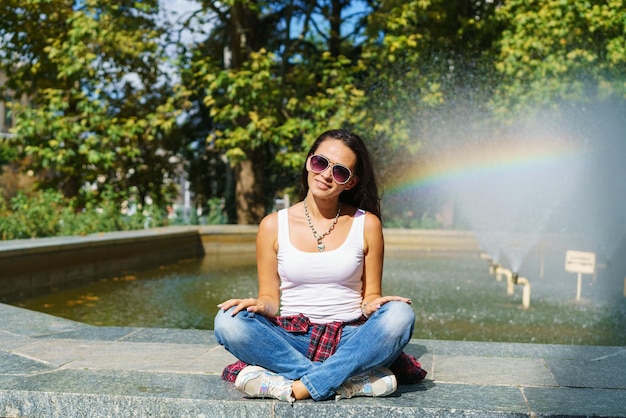 Vrolijke jonge vrouw in zonnebril zit tegen achtergrondfontein met regenboog