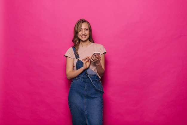 Vrolijke jonge vrouw met behulp van mobiele telefoon en kijken naar de camera op geïsoleerde roze achtergrond. Gelukkig mooi meisje in denim kleding poseren in professionele studio.