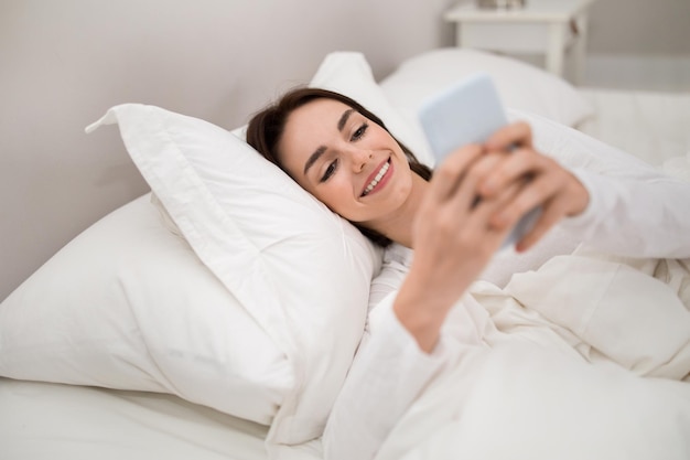 Vrolijke jonge vrouw met behulp van telefoon in bed