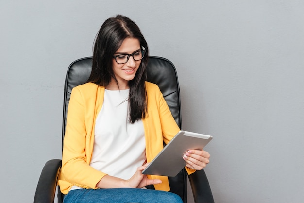 Vrolijke jonge vrouw met een bril en gekleed in een gele jas zittend op een bureaustoel tijdens het gebruik van een tabletcomputer over een grijs oppervlak.