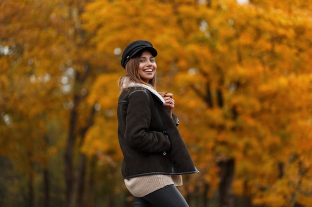 Vrolijke jonge vrouw met een positieve glimlach in een jas in een trendy hoed met een leren tas poseren in herfst park op een achtergrond van bomen met oranje gebladerte. Blij meisje in het bos. Casual uitstraling.