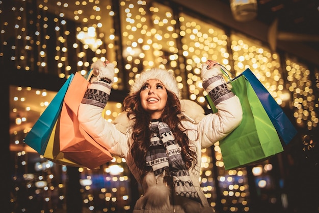 Vrolijke jonge vrouw met kleurrijke boodschappentassen die plezier heeft in de straat van de stad met Kerstmis.