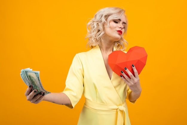 Vrolijke jonge vrouw met kort blond haar glimlacht en kiest tussen liefde en geld geïsoleerd op oranje