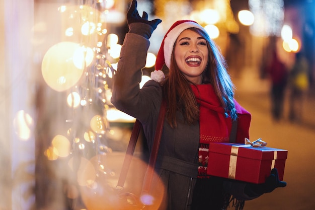 Vrolijke jonge vrouw met rode huidige doos heeft plezier in de stadsstraat met Kerstmis.