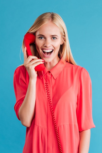 Vrolijke jonge vrouw praten over rode telefoon en lachen geïsoleerd op een blauwe muur