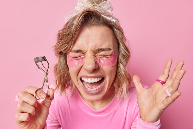 Foto vrolijke jonge vrouw roept luid houdt mond open houdt wimpers krultang doet make-up houdt hand omhoog brengt hydrogelpleisters onder de ogen aan voor het gladmaken van de huid geïsoleerd op roze achtergrond