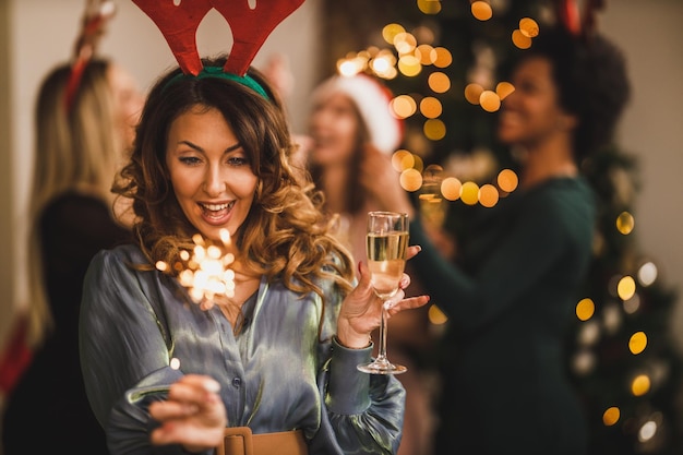Vrolijke jonge vrouw roostert met champagne en speelt met sterretjes terwijl ze Kerstmis of Nieuwjaar viert met haar vrienden op een thuisfeest.