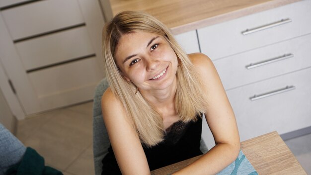 Vrolijke jonge vrouw zit aan tafel in een gezellige ingerichte kamer terwijl ze lacht en naar de camera kijkt