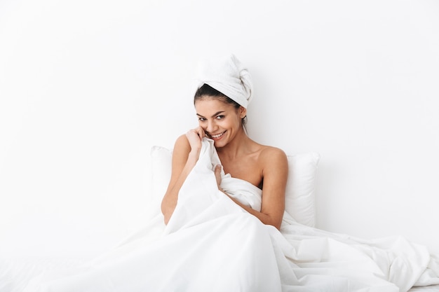 Vrolijke jonge vrouw zittend in bed na het douchen gewikkeld in deken, poseren