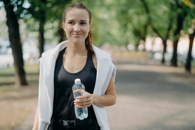 Vrolijke jonge vrouwelijke hardloper houdt waterfles poses buiten in actieve slijtage neemt pauze na training wandelingen in het park krijgt verfrissing na het joggen voelt dorstig blijft fit met regelmatige sport