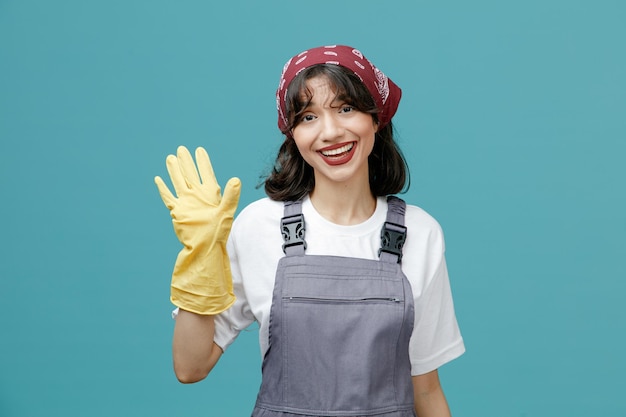 Vrolijke jonge vrouwelijke schoonmaker die uniforme bandana en rubberen handschoenen draagt en naar de camera kijkt die hallo gebaar toont geïsoleerd op blauwe achtergrond