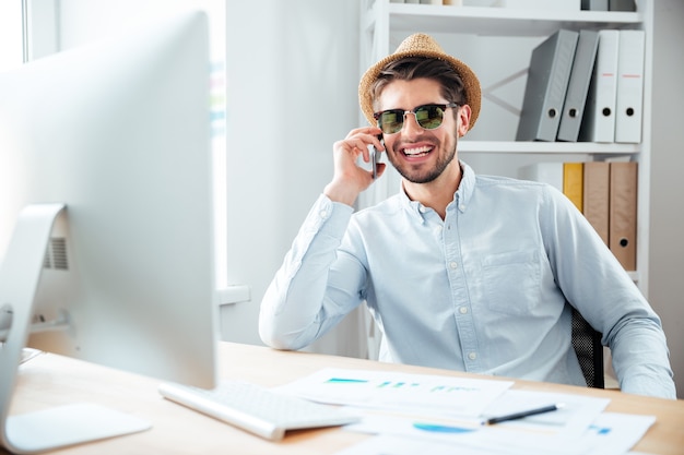 Vrolijke jonge zakenman in hoed en zonnebril werken met computer en praten om mobiele telefoon in office