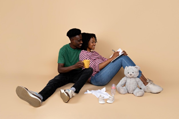 Vrolijke jonge zwarte man en dame met dikke buik zitten op de vloer genieten van zwangerschap met speelgoed drinken thee