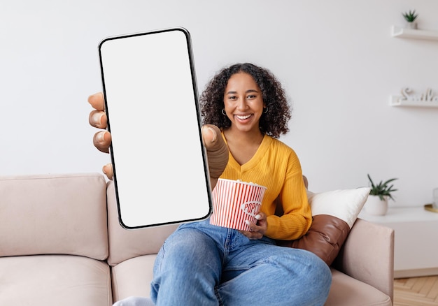 Vrolijke jonge zwarte vrouw die popcorn vasthoudt en een grote lege smartphone toont