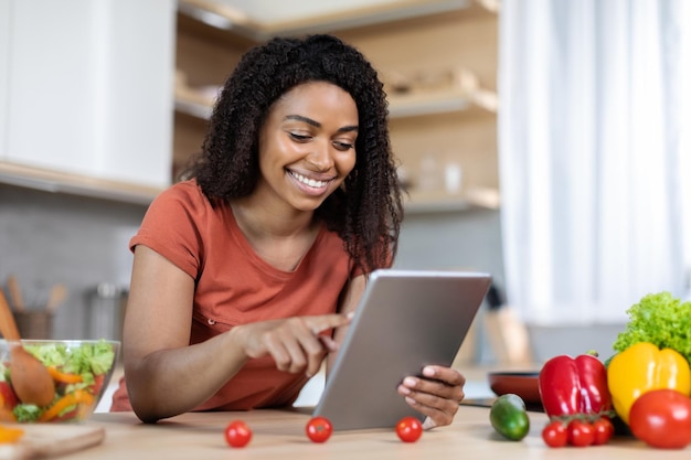 Vrolijke jonge zwarte vrouw in rode tshirt typen op tablet lees bericht aan tafel met biologische groenten