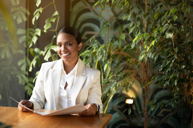 Vrolijke jonge zwarte vrouw in wit pak zittend aan tafel leesmenu in eco café met planten