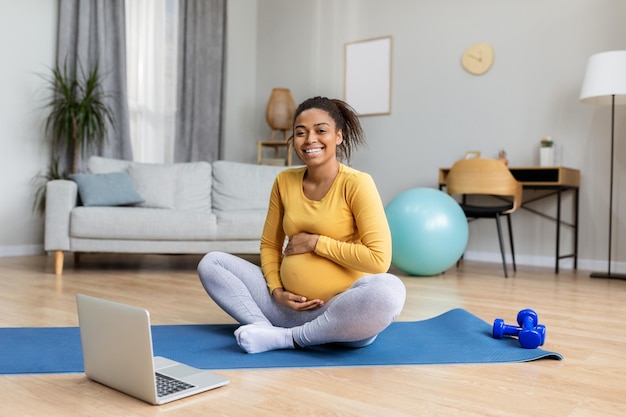 Vrolijke jonge zwarte zwangere vrouw raakt grote buik aan die yoga beoefent op mat bekijk trainingsvideo op laptop