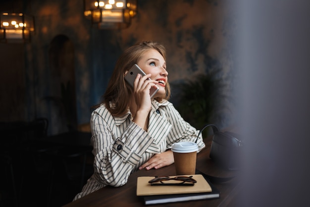 Vrolijke jongedame in gestreepte trenchcoat gelukkig praten over mobiel