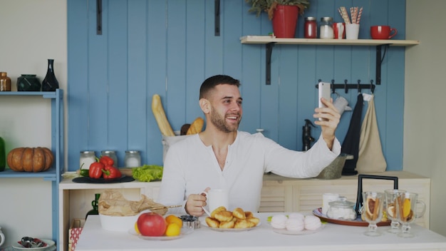 Vrolijke jongeman die smartphone gebruikt voor online videochat met vriendin tijdens het ontbijt