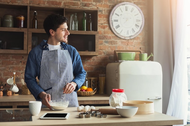 Vrolijke jongeman die taart bakt in de loftkeuken thuis, met behulp van digitale tablet met recept, kopieer ruimte