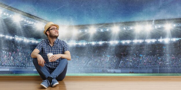 Foto vrolijke jongeman in casual kleding en hoed zittend op de vloer, stadion achtergrond. gemengde media