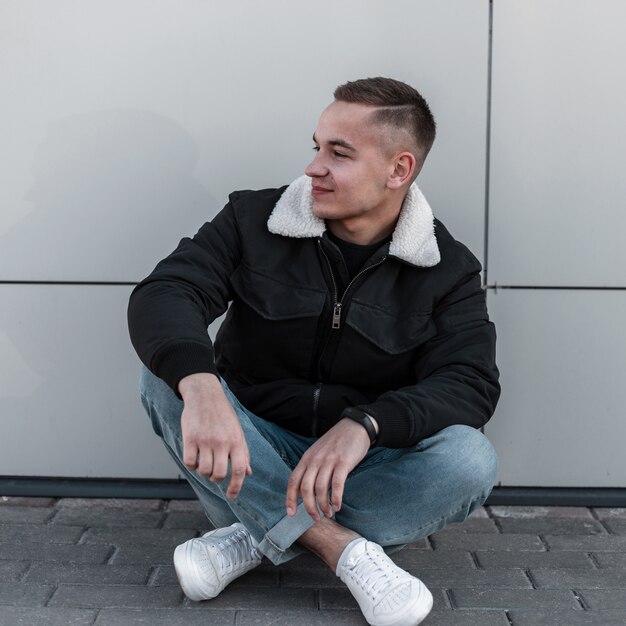 Vrolijke jongeman in modieuze lente kleding in lederen witte sneakers poseren zittend op de stoep in de buurt van een modern gebouw in de stad in de lente