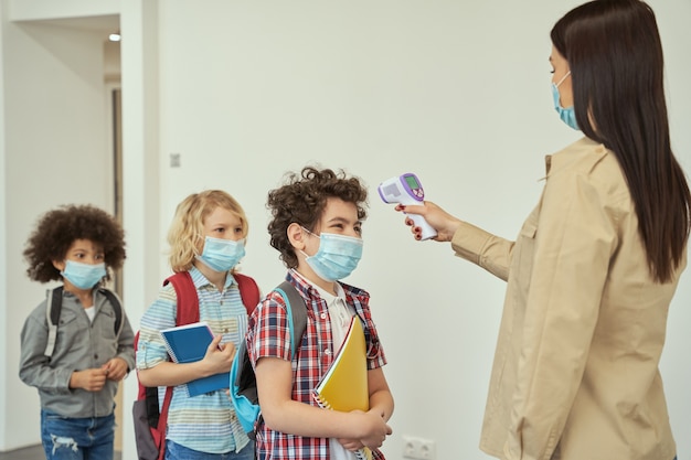 Vrolijke jongen die een gezichtsmasker draagt, kijkt naar zijn leraar die de temperatuur van het kind meet met