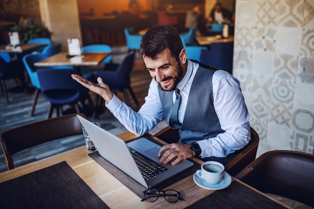 Vrolijke Kaukasische bebaarde knappe zakenman zitten in café en met behulp van laptop. Op tafel staan laptop, koffie, bril en water.