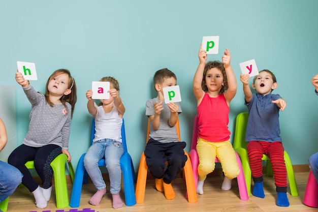 Vrolijke kinderen tijdens voorschoolse les Betrokken kinderen zitten op kleurrijke stoelen met kaarten met letters die hen aan de camera tonen Communicatieve onderwijsbenadering