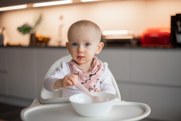 Vrolijke kleine baby eet alleen met een lepel terwijl hij in een kinderstoel zit