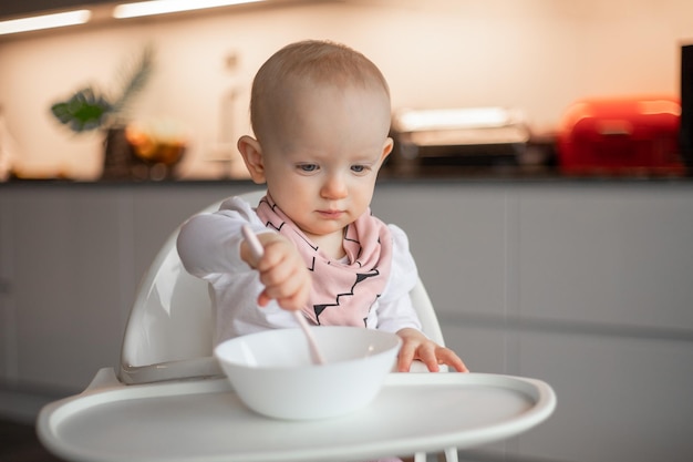 Vrolijke kleine baby eet alleen met een lepel terwijl hij in een kinderstoel zit