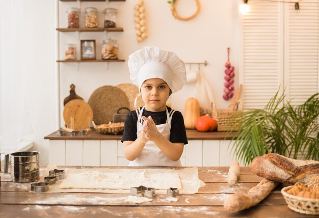 Vrolijke kleine chef-kok in een pak zit aan een houten tafel en maakt brood van deeg