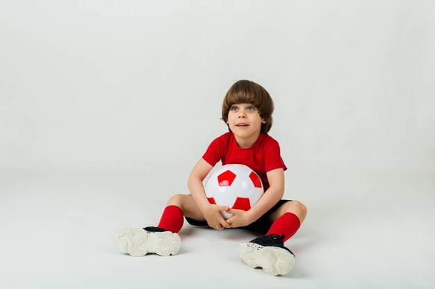 Vrolijke kleine jongen in uniform zit met een voetbal op een wit oppervlak met ruimte voor tekst