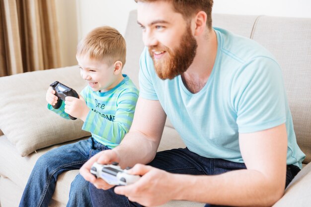 Vrolijke kleine zoon en vader die thuis samen computerspelletjes spelen en glimlachen