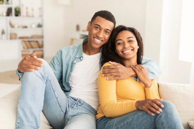 Vrolijke knappe jonge afro-amerikaanse man die zijn vrouw knuffelt terwijl ze op de bank rust, geniet van het weekend