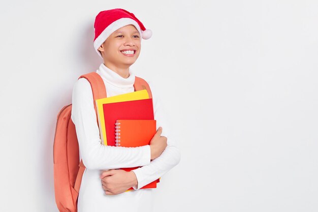 Vrolijke knappe jonge Aziatische student in een kerstmuts met t-shirt met rugzak met boek geïsoleerd op een witte achtergrond