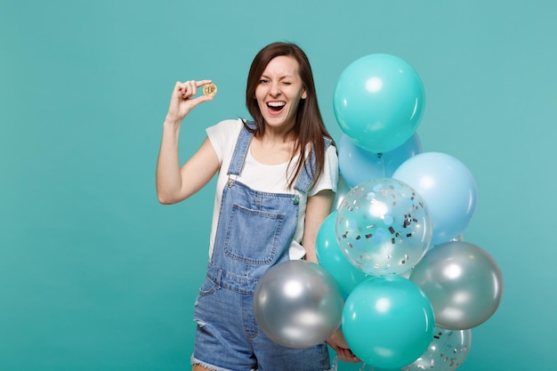 Vrolijke knipperende jonge vrouw met bitcoin, toekomstige valuta en vieren met kleurrijke luchtballonnen geïsoleerd op blauwe turquoise muur achtergrond. Vakantie verjaardagsfeestje, mensen emoties concept.
