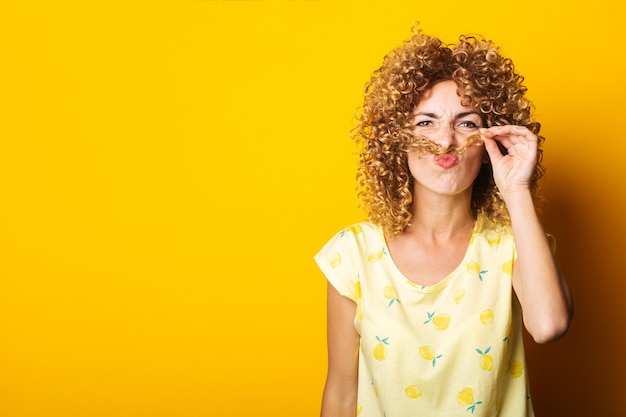 Vrolijke krullende jonge vrouw die snor met haar op gele achtergrond doet.