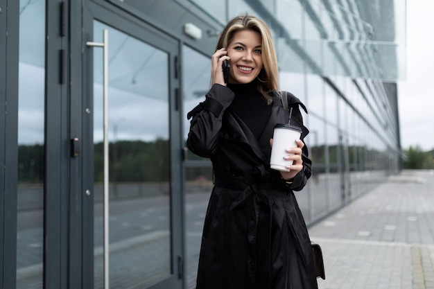 Vrolijke lachende caucasiaanse vrouw in zwarte zakelijke kleding die aan de telefoon praat met een kopje