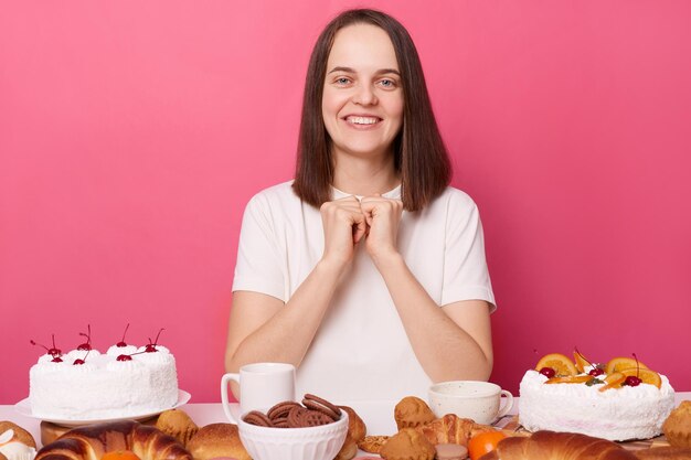 Vrolijke lachende opgetogen bruinharige vrouw in wit t-shirt zittend aan tafel met heerlijke desserts geïsoleerd op roze achtergrond kijkend naar camera brekend dieet