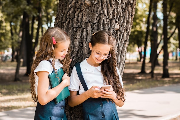 Vrolijke leerlingen in schooluniform met een boek kijken naar een smartphone in het park op een warme dag