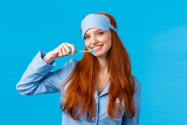 Vrolijke leuke roodharige vrouw in nachtkleding, pyjama en slaapmasker, tanden poetsen met tandenborstel en vrolijk lachend, wakker worden en hygiëne verzorgen, blauwe muur staan