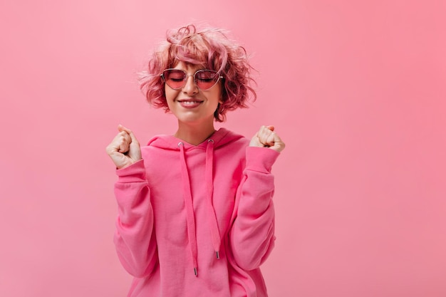 Vrolijke leuke vrouw in roze heldere hoodie doet wens en poseert met geklede ogen Pinkhaired meisje in zonnebril poses op geïsoleerde achtergrond