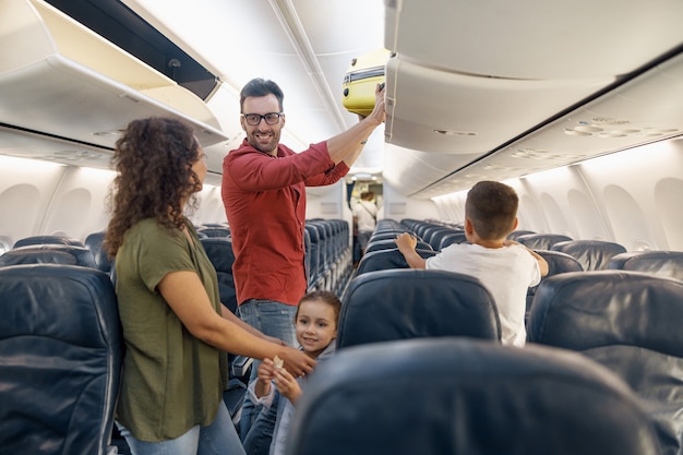 Vrolijke man die met een glimlach naar zijn vrouw kijkt en bagage in het compartiment zet terwijl hij samen met zijn gezin per vliegtuig reist. Familievakantie, vervoersconcept