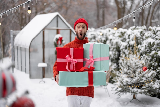 Vrolijke man in rode trui en hoed draagt cadeau dozen in de besneeuwde achtertuin Concept van gelukkige wintertijd Dankbaarheidsdag en Valentijnsdag