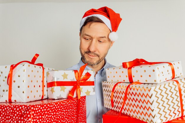 Vrolijke man in t-shirt in rode kerstmuts met veel geschenkdozen die tegen een grijze achtergrond staan, studio opname. Zakenman is klaar voor Kerstmis in zijn kantoor