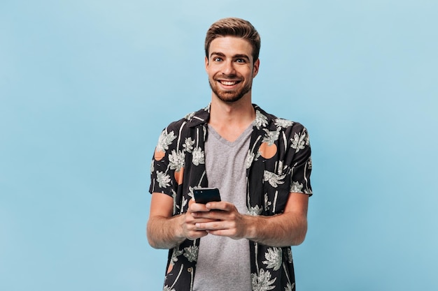 Vrolijke man met gemberbaard in cool zwart bedrukt shirt en geruite t-shirt die in de camera kijkt terwijl hij glimlacht en de telefoon vasthoudt