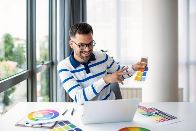 Vrolijke man-ontwerper die videoconferentie heeft met klanten die aan een bureau achter een computer zitten met kleurenpaletten gebaren en glimlachende kopieerruimte